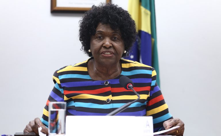 Deputada Benedita da Silva fala ao microfone. Ela é negra, tem cabelo curto e usa uma blusa com listras coloridas