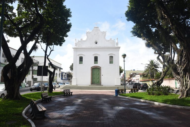 Uma igreja branca numa praça com árvores num dia de sol.